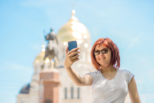 Beautiful smiling tourist woman holding smartphone and taking photo on city square or street in sunny day. Vacation and travel concept