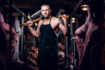 Shirtless tattooed butcher in apron posing with ax in a refrigerated warehouse in the midst of meat carcasses