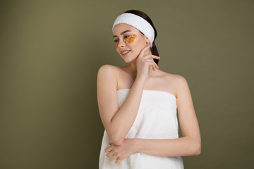 Young woman wrapped in towel on green background applied gold patches under eyes