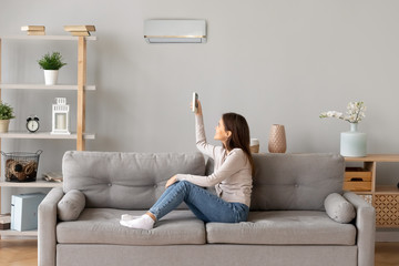 Young smiling woman sitting on couch switching on air conditioner
