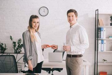 Obraz premium happy businessman holding pills and glass of water near cheerful blonde colleague in office