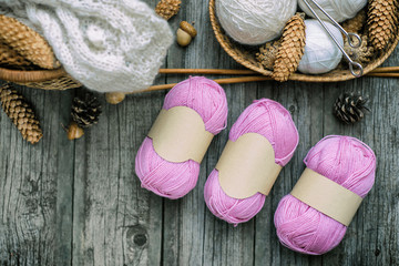 Three pink balls of wool threads with empty labels lying on vintage wood desk with knitting needles and basket.