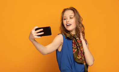 Fototapeta premium Selfie time! Attractive young woman in casual wear making selfie by her smart phone and smiling while standing against yellow background