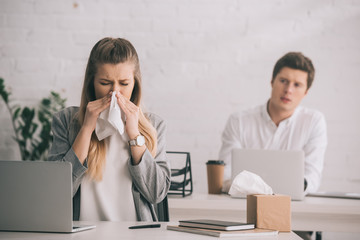 blonde businesswoman sneezing in tissue near coworker in office