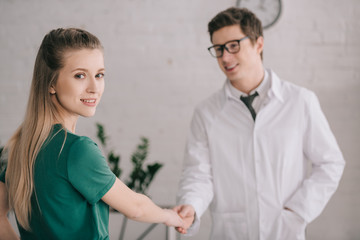 selective focus of cheerful blonde patient shaking hands with happy doctor in white coat