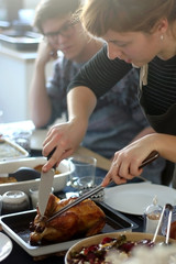 Young woman cutting roast chicken at lunch. Male friend siting in the background. Selective focus. 