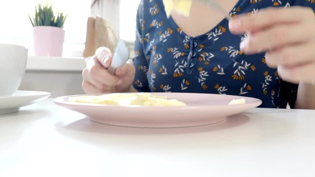 Woman sitting by the table, eating an omlet with a folk and a knife and drinking coffee or tea for breakfast at home or restaurant