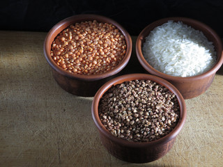 Grains of buckwheat, lentils and rice in clay bowls on a board, against a dark background