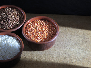 Grains of buckwheat, lentils and rice in clay bowls on a board, against a dark background