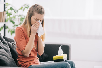 attractive blonde woman sneezing while sitting on sofa with tissue box