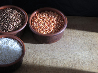 Grains of buckwheat, lentils and rice in clay bowls on a board, against a dark background