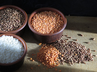 Grains of buckwheat, lentils and rice in clay bowls on a board, against a dark background