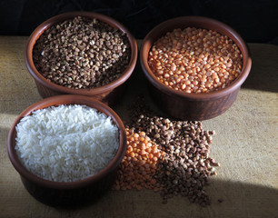 Grains of buckwheat, lentils and rice in clay bowls on a board, against a dark background