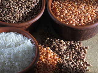 Grains of buckwheat, lentils and rice in clay bowls on a board, against a dark background