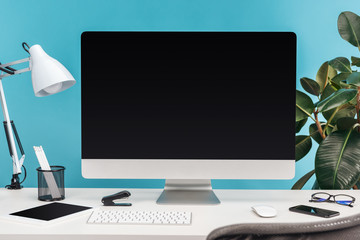 workplace with gadgets, lamp, stationery on white table near green plant on blue background
