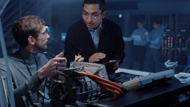 Two Professional Automotive Engineers with a Tablet Computer and Inspection Tools are Having a Conversation While Testing an Electric Engine in a High Tech Laboratory with a Concept Car Chassis.