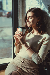 Young attractive girl sitting alone near big window in coffee shop during free time and working. Sunny day