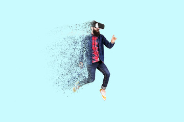 Fototapeta na wymiar Full length portrait of a jumping bearded hipster in glasses of virtual reality, man dispersing and disintegrating into particles ,concept of new technologies