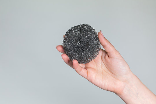 Close Up View Of One Female Hand Holding New Round Metal Sponge For Washing Dirty Frying Pans And Saucepans Isolated At Grey Background. Horizontal Color Photography.