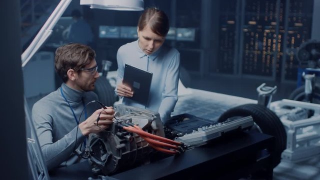 Male and Female Automotive Engineers with a Tablet Computer and Inspection Tools are Having a Conversation While Testing an Electric Engine in a High Tech Laboratory with a Concept Car Chassis.