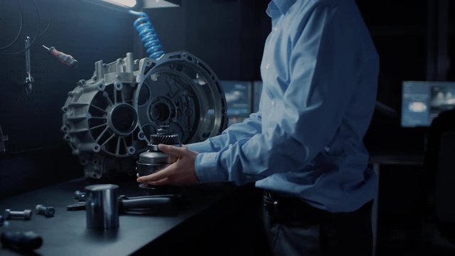 Professional Automotive Engineer in Glasses is Working on Transmission Gears in a High Tech Innovative Laboratory with a Computer Screens.