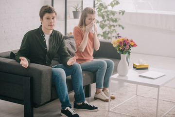 Obraz premium handsome man showing shrug gesture while sitting near vase with flowers and sneezing blonde girl with pollen allergy