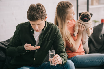selective focus of handsome man allergic to dog holding glass of water and looking at pills near blonde woman with pug