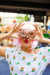 bright fashion funny girl in pink wig posing on background carousel at an amusement park. The hands holding the sweet donuts