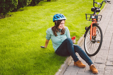Subject ecological mode of transport bicycle. Beautiful young Caucasian woman wearing a blue helmet and long hair poses standing next to an orange-colored rental bike with a basket in a city park