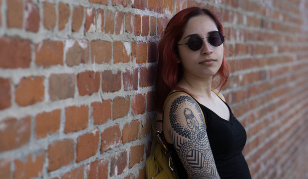 portrait of hip young tattooed woman against bricks wearing sunglasses
