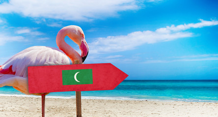 Maldives flag on wooden table sign on beach background with pink flamingo. There is beach and clear water of sea and blue sky in the background. It is tropical natural background.
