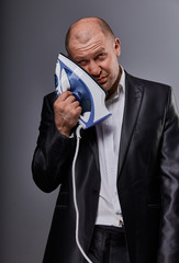 Bald loser suffering business man holding the home iron near the face with sad face in suit on grey...