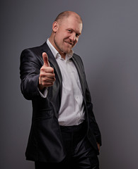 Exited comic bald business man in black suit showing the finger success thumb up sign on grey background. Closeup
