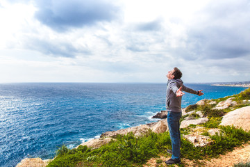 Rear view of a young guy standing on a seaside rock cliff edge with arms spread open against a seascape