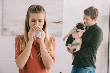 selective focus of blonde girl allergic to dog sneezing in white tissue near man with cute pet