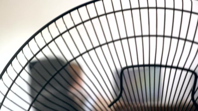 close-up of part of an office fan, on the background of a blurred writing desk, at which a person works at a computer