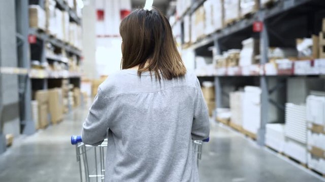 Young Asian woman rides shopping cart choosing new furniture in warehouse, teenager female feeling happy in front of shelves in department store. Women shopping new furniture concept.