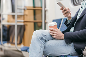 Unrecognizable man in blue coat is sitting in a subway train, drinking coffee and surfing in web with his smartphone. Efficient use of time on the way to work. People and communication concept.