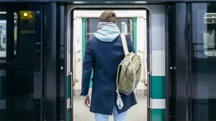 Fototapeta na wymiar Close up rear view of handsome man in blue coat and green backpack behind stands in front of the open doors of the subway and waiting for next train. Way to work. Urban life concept.