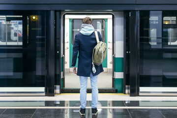 Rear view of handsome man in blue coat and green backpack behind stands in front of the open doors...