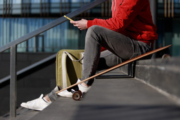 Stylish man longboarder in casual clothes using his smartphone, resting on the steps, sitting with longboard/skateboard outdoors, top view, cropped image. Urban, subculture, skateboarding concept