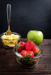 Small glass bowls of fresh strawberries, pineapple chunks and a whole green apple served on wooden board with a dark background
