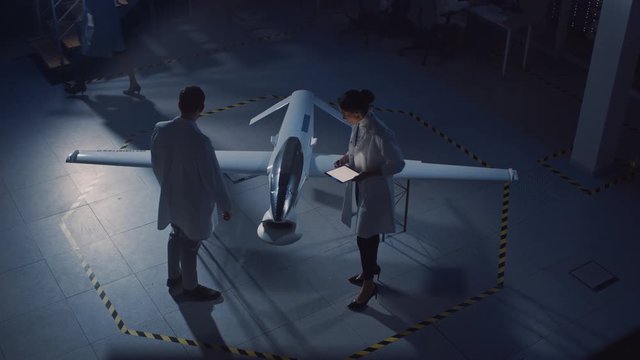 Two Aerospace Engineers Work On Unmanned Aerial Vehicle / Drone Prototype. Aviation Scientists in White Coats Talking, Using Tablet Computer. Industrial Laboratory with Surveillance. Elevated Shot