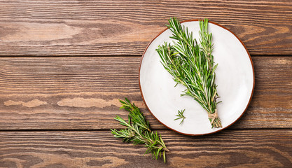 Sprigs of fresh fragrant rosemary are collected in a bun lying on a white plate on a wooden background. Copy space.