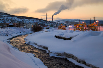 Evening urban landscape with a small river, buildings, hills and fuming pipes. On the banks of the river playground. Twilight after sunset. Magadanka River. Magadan, Siberia, Far East of Russia.