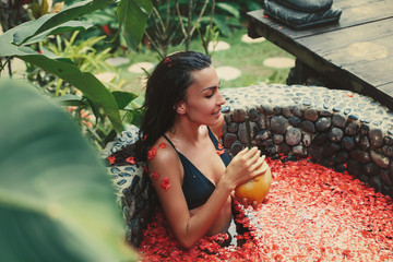 Woman wearing black swimming suit relaxing in tropical open air spa bath and drink coconut