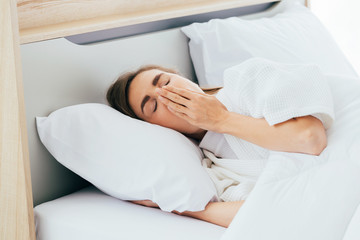 Fototapeta na wymiar Sleepy Caucasian female with closed eyes covering mouth with hand and yawning while lying on comfortable bed in morning