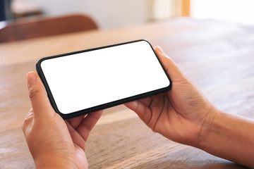 Mockup image of woman's hands holding black mobile phone with blank white screen horizontally on wooden table