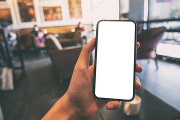 Mockup image of a hand holding and showing black mobile phone with blank white screen in cafe