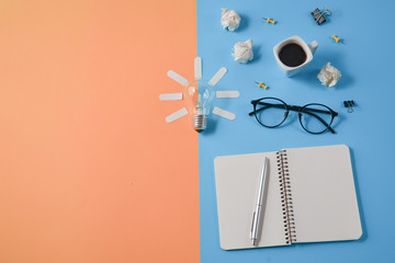 Flat lay financial planning brainstorming messy table top image with blank clip board, office supplies, pen, notepad, eyeglasses, coffee cup, light bulb on orange and blue background.